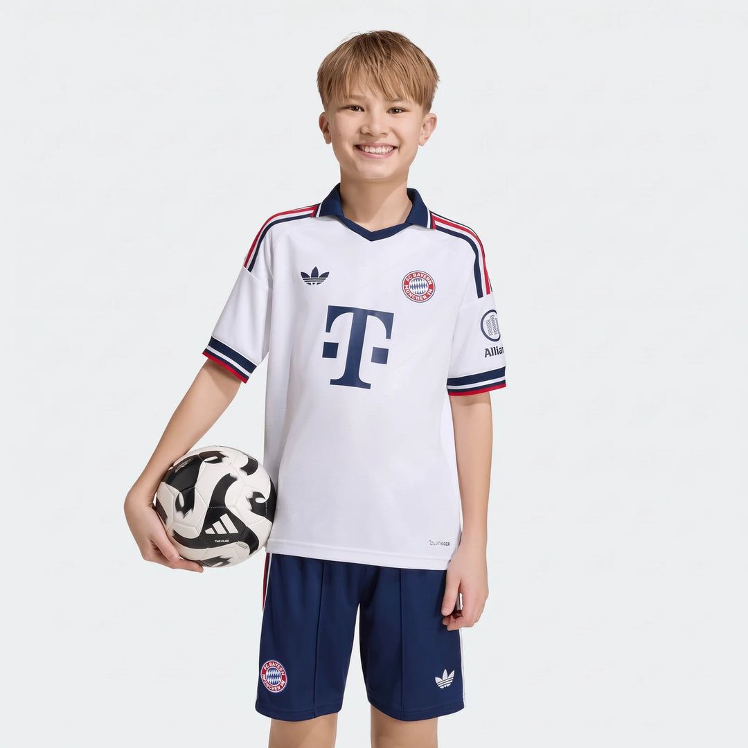 A young boy smiles while wearing the white Adidas Fc Bayern 26/27 Away Jersey for kids and blue shorts, holding a soccer