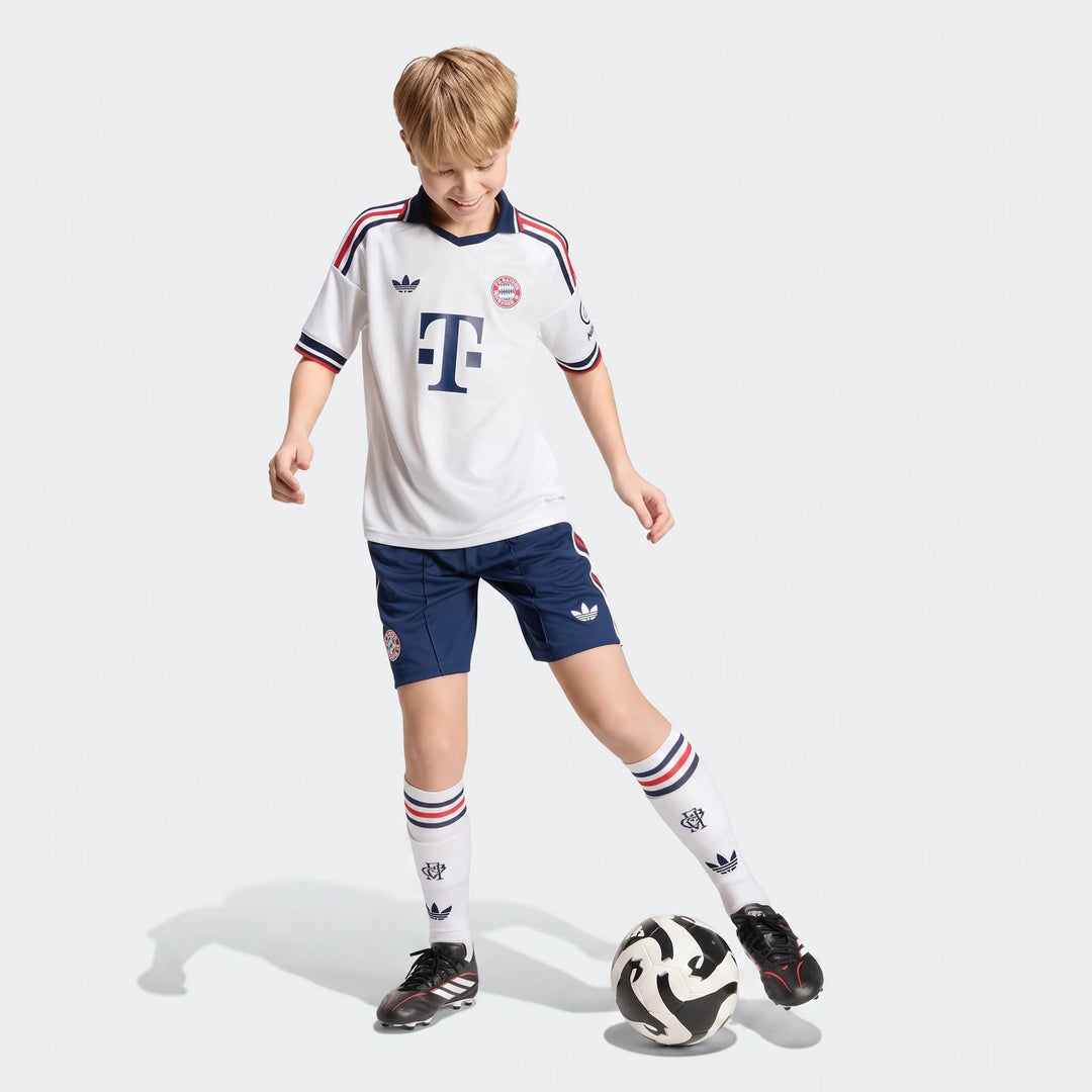 Young boy wearing Fc Bayern 26/27 Away Jersey Kids, blue shorts, and white socks, dribbling a soccer ball in a studio