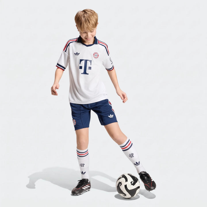 Young boy wearing Fc Bayern 26/27 Away Jersey Kids, blue shorts, and white socks, dribbling a soccer ball in a studio