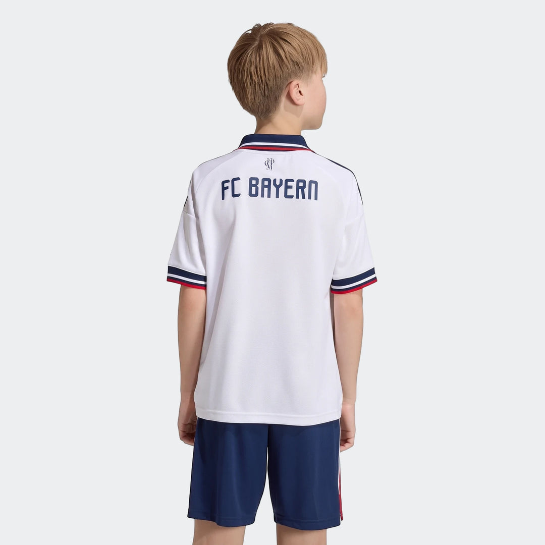 Young boy wearing white FC Bayern 26/27 Away Jersey and blue shorts, back view, showcasing the team name and striped details.