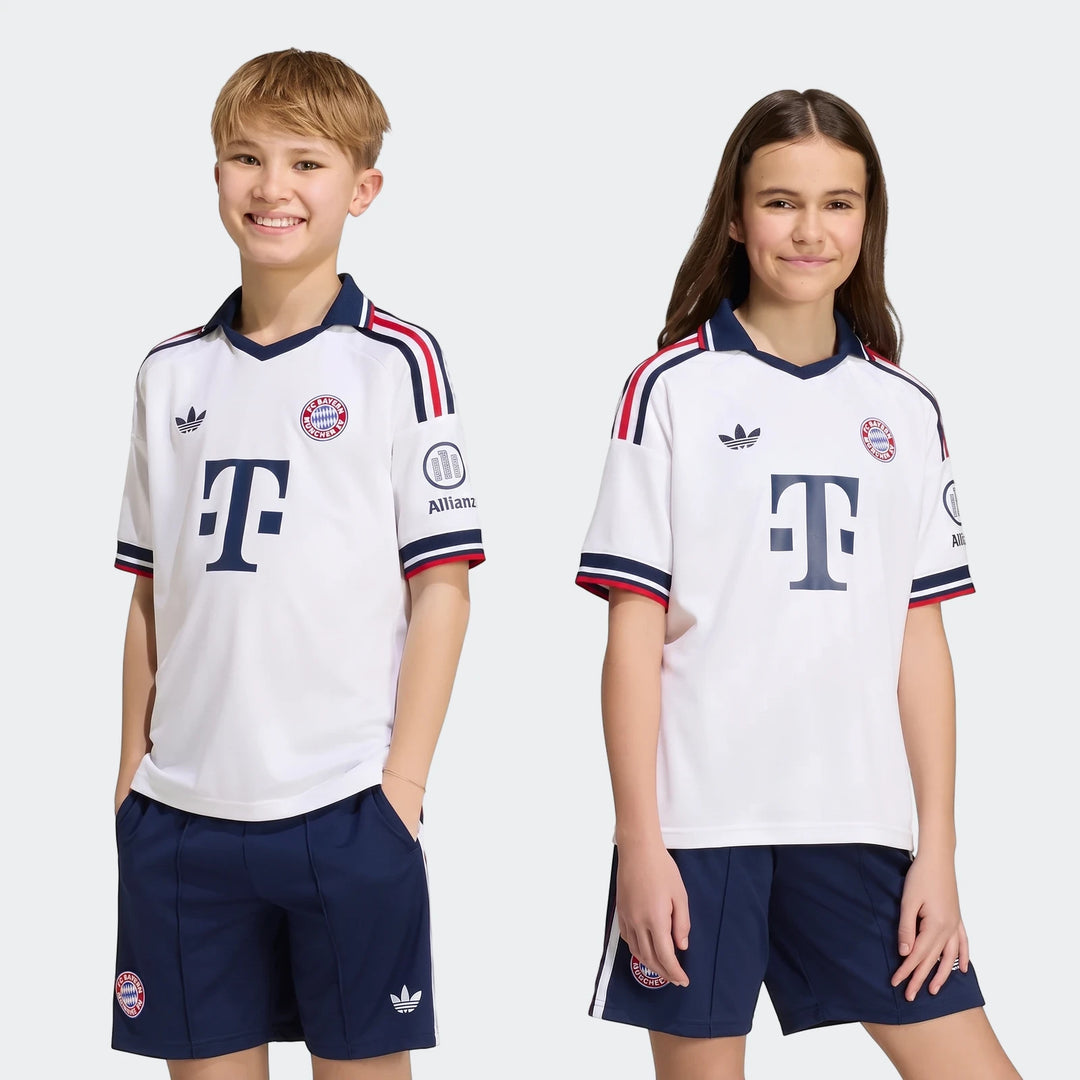 Two children smile, modeling the white Fc Bayern 26/27 Away Jersey and navy shorts with team crests and Adidas logos.