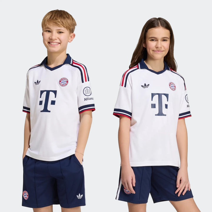 Two children smile, modeling the white Fc Bayern 26/27 Away Jersey and navy shorts with team crests and Adidas logos.
