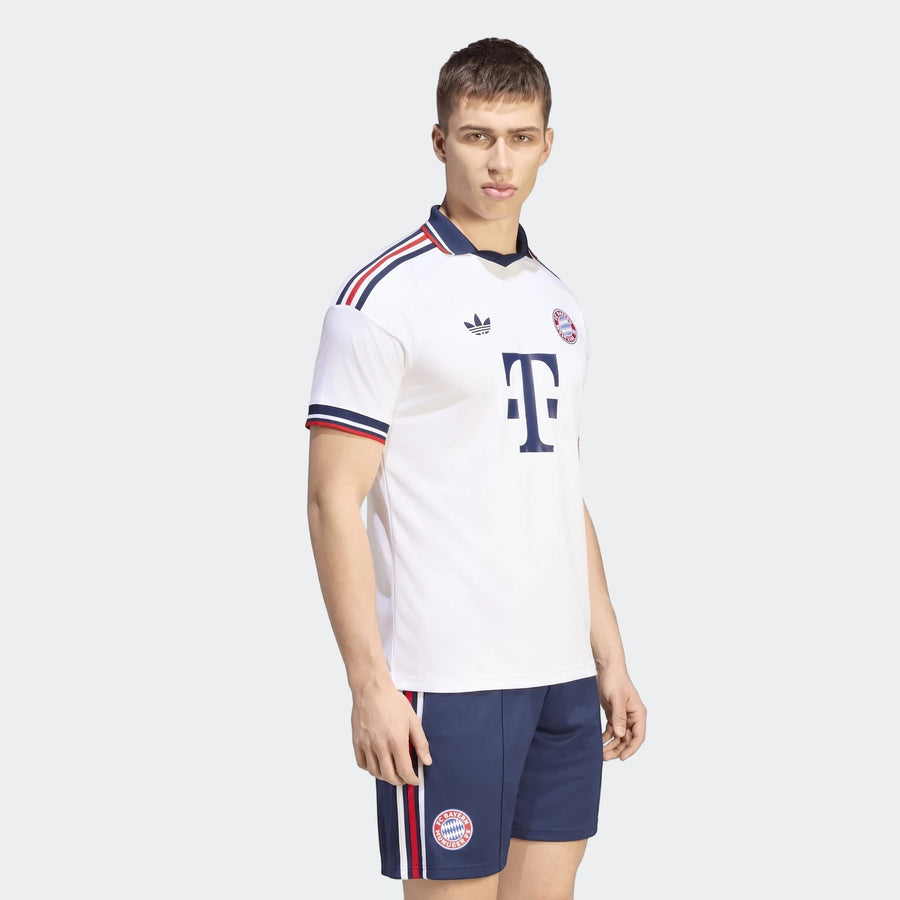 Male model wearing a white Fc Bayern 26/27 Away Jersey with navy shorts, featuring striped details and club crests.