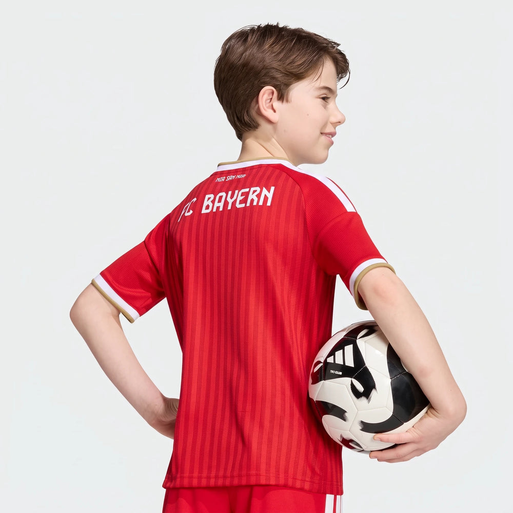 Young boy from behind wearing a red Adidas FC Bayern 26/27 Home Jersey for kids, holding a black and white soccer ball.