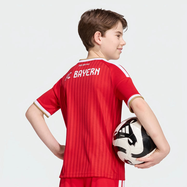 Young boy from behind wearing a red Adidas FC Bayern 26/27 Home Jersey for kids, holding a black and white soccer ball.