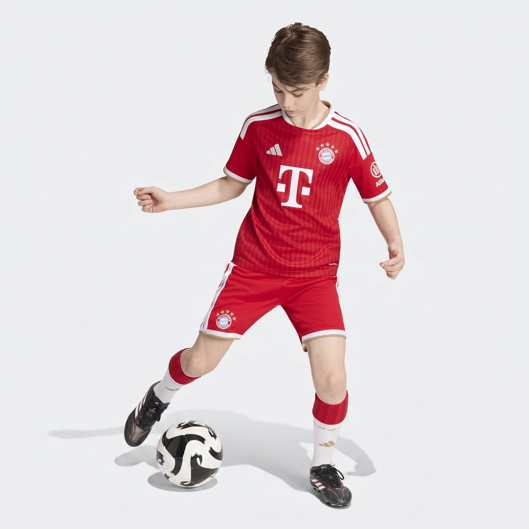 Red Adidas FC Bayern 26/27 Home Jersey for kids, worn by a young boy actively kicking a soccer ball.