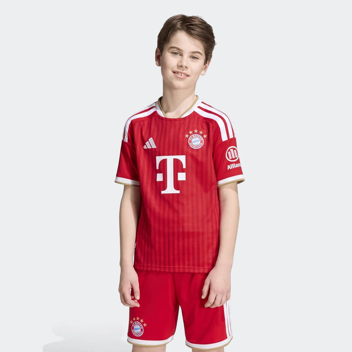 Young boy wearing a red Adidas FC Bayern 26/27 Home Jersey and matching shorts, front view.