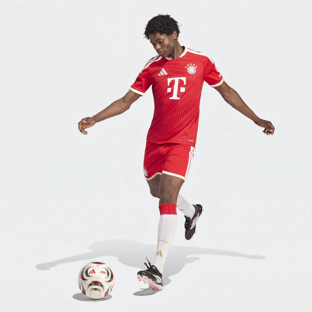 Male model wearing a red and white Adidas FC Bayern 26/27 Home Jersey, red shorts, and white socks, kicking a football.