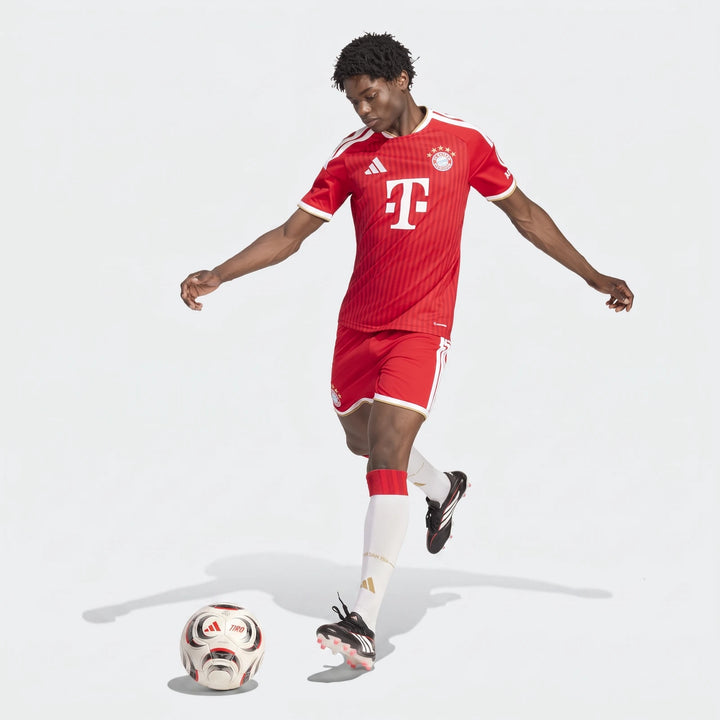 Male model wearing a red and white Adidas FC Bayern 26/27 Home Jersey, red shorts, and white socks, kicking a football.