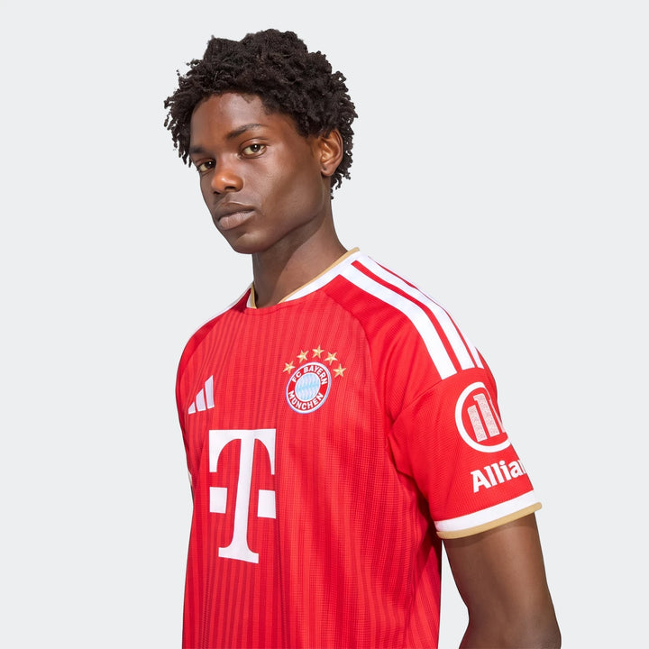 Close-up of a male model wearing the red Fc Bayern 26/27 Home Jersey with white stripes on shoulders and team logos.