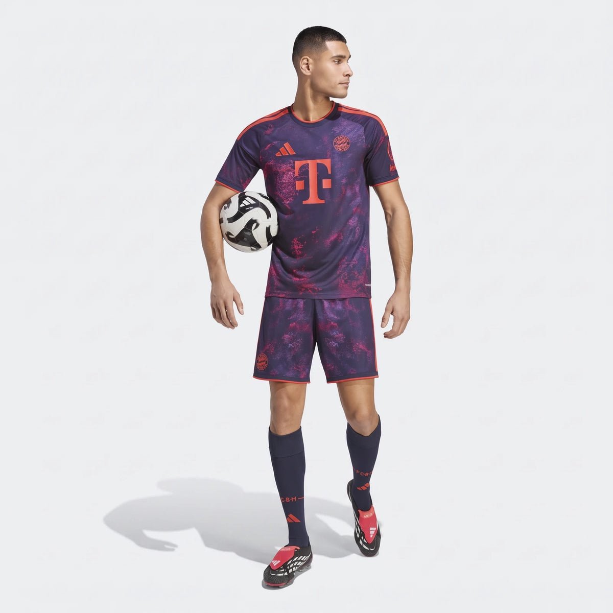 Male model in a full FC Bayern 26/27 Third Jersey kit, featuring a purple and orange design, holding a soccer ball.