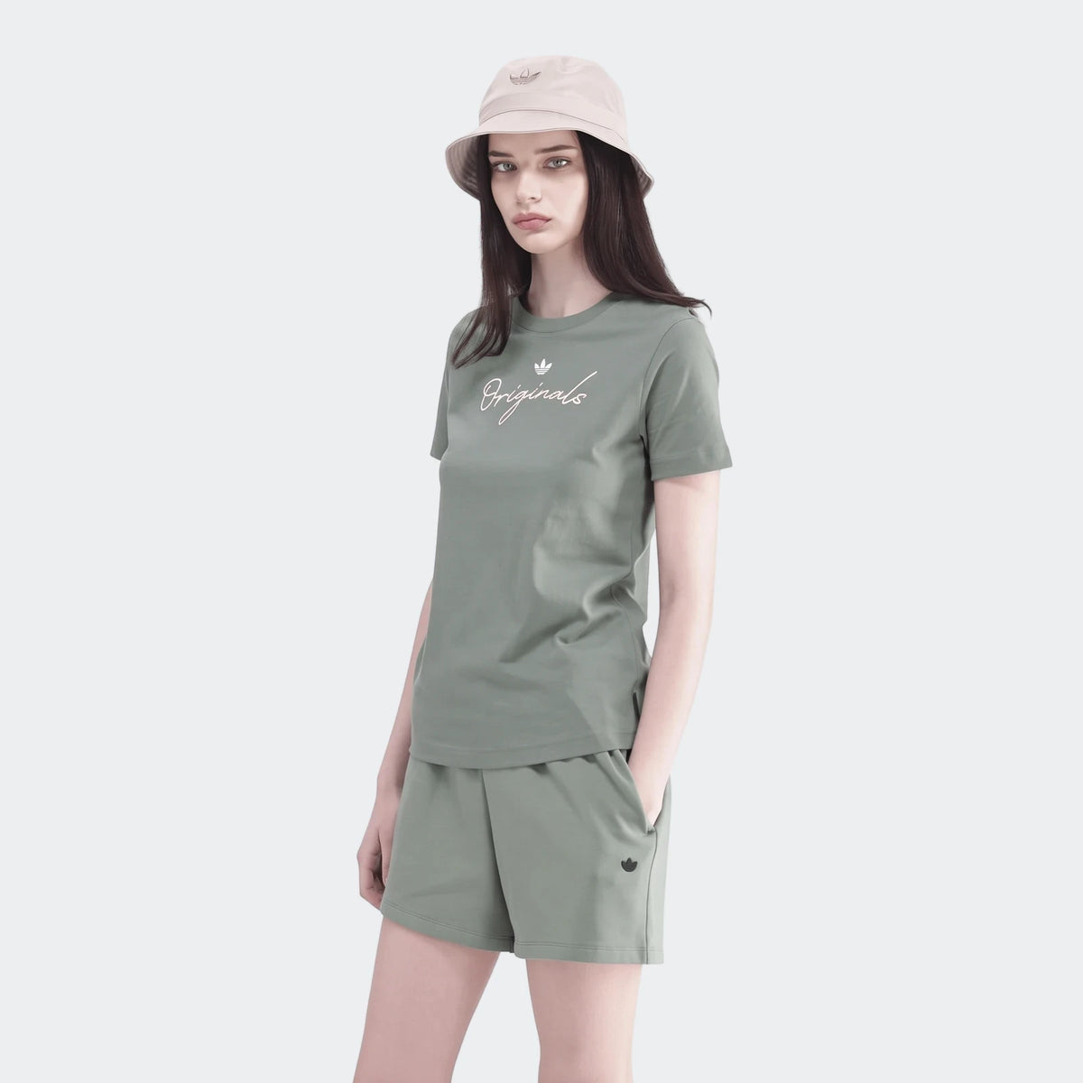 Female model wearing a greenish-grey Adidas Originals short sleeve t-shirt, matching shorts, and a beige bucket hat.