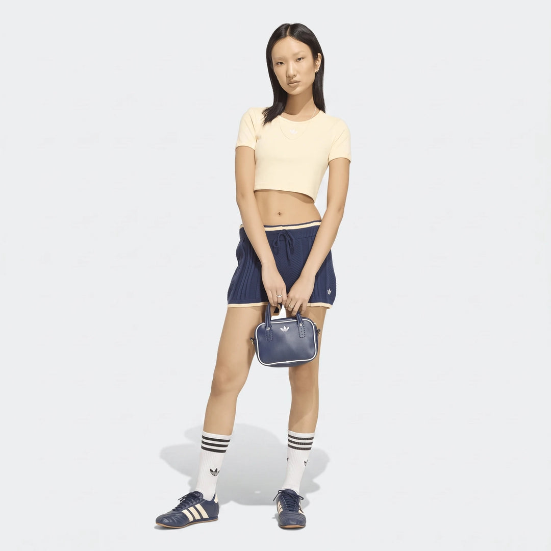 A female model wearing navy blue cable knit shorts and a cream crop top, posing with a small matching handbag and sneakers.