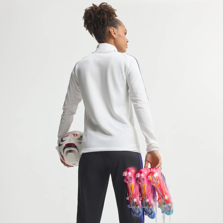 Female soccer player in a white jacket and black pants, holding F50 Hyperfast Elite Firm Ground Football Boots and a soccer