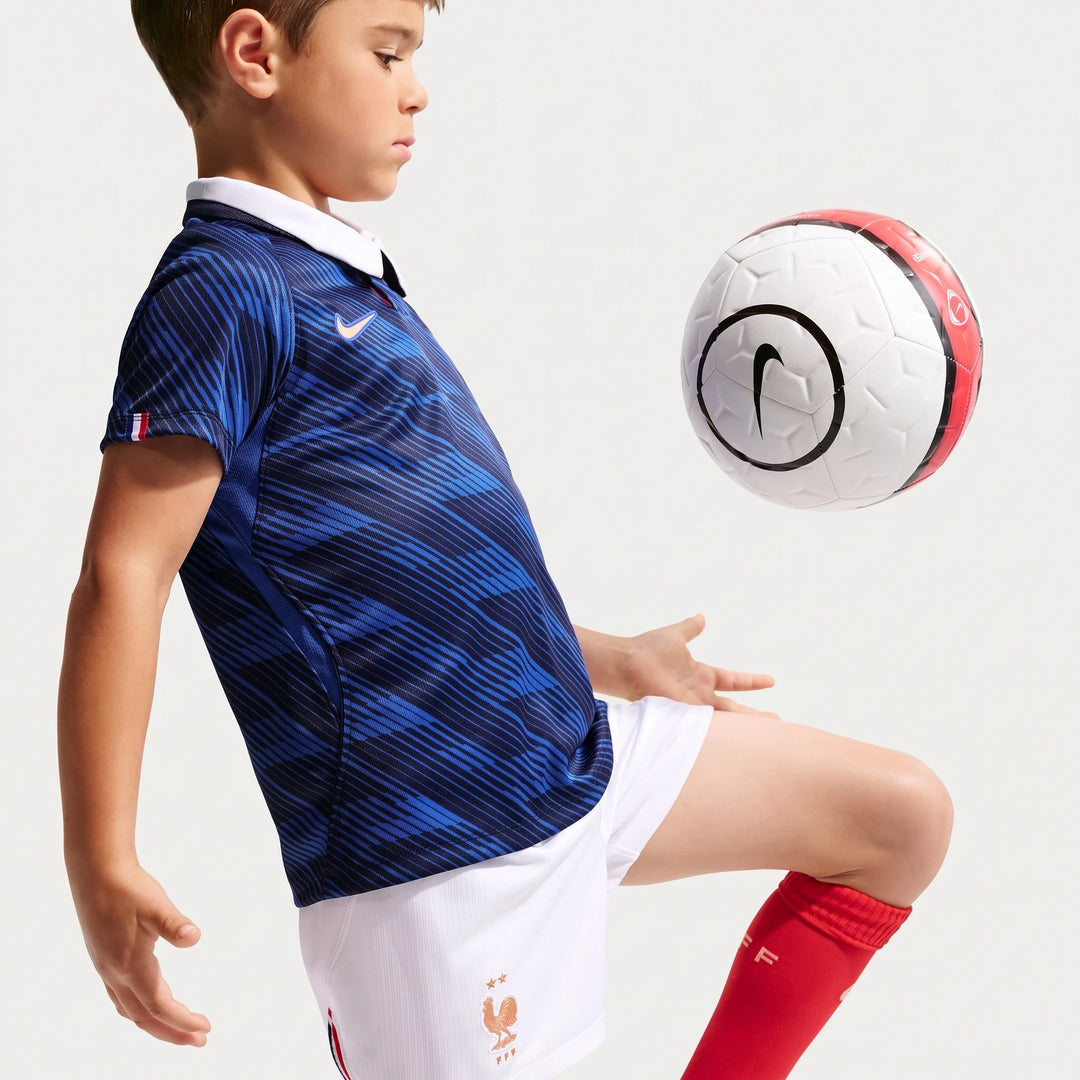 Young boy in a blue Fff 2026 Stadium Home Nike Soccer Replica 3-Piece Kit, kicking a white soccer ball.