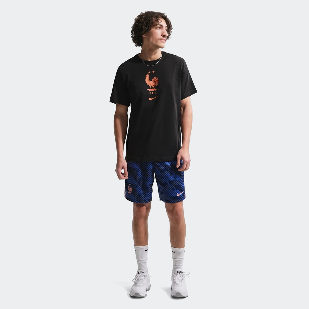 A man wears an Fff Black Nike Soccer T-Shirt with blue patterned shorts, white socks, and white sneakers, standing