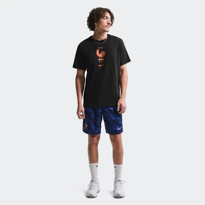 A man wears an Fff Black Nike Soccer T-Shirt with blue patterned shorts, white socks, and white sneakers, standing