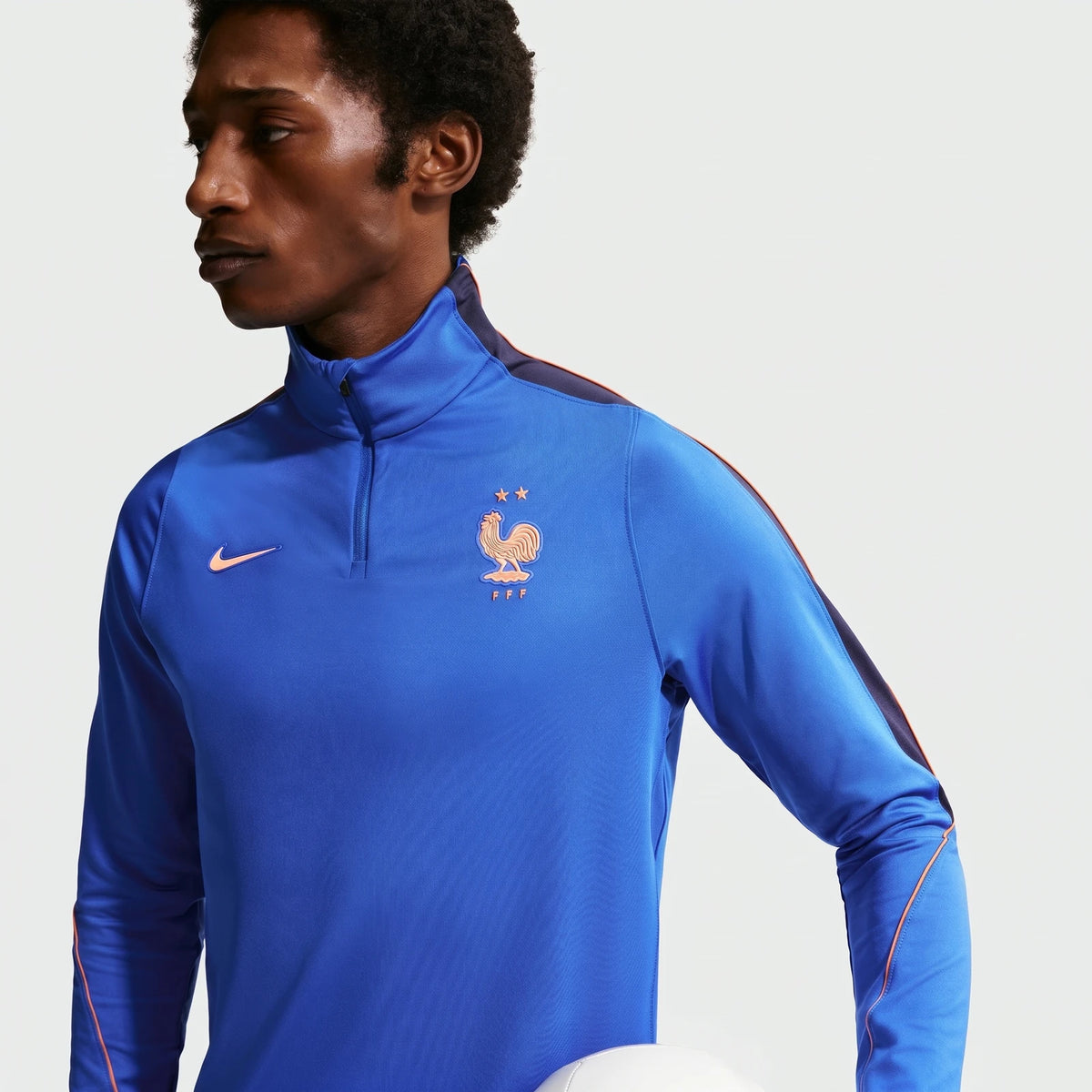 A male model wears a blue Nike Dri-Fit FFF Strike soccer drill top with orange accents and quarter-zip, holding a white