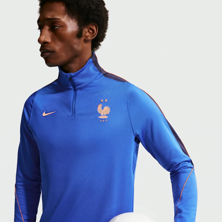 A male model wears a blue Nike Dri-Fit FFF Strike soccer drill top with orange accents and quarter-zip, holding a white