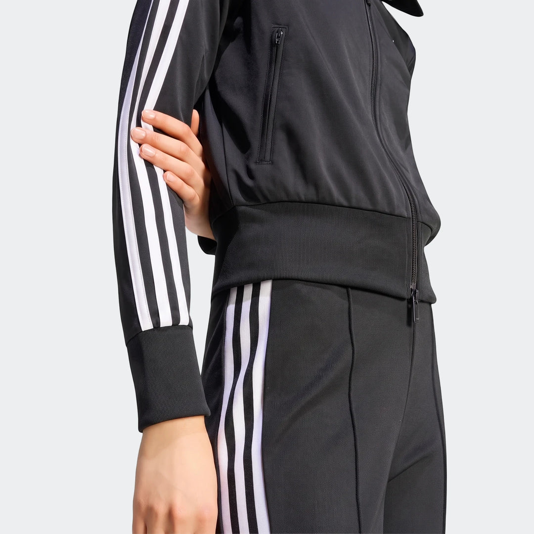 Close-up of a person wearing a black Adidas Firebird Classic Track Jacket with white three-stripe detailing on the sleeves