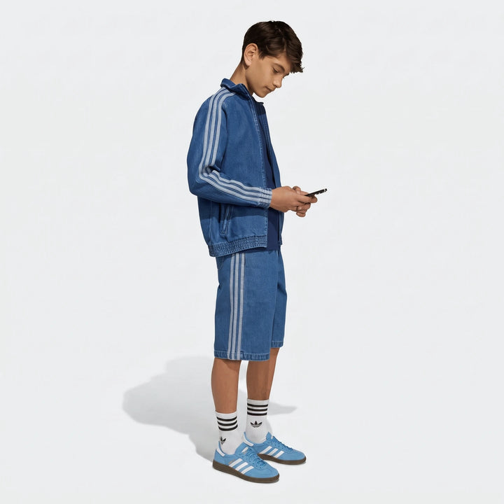 Side view of a young man wearing a blue denim Firebird Track Top with matching shorts, white stripes, and light blue