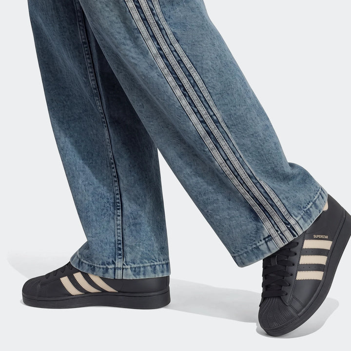 Close-up of a person wearing blue Firebird Loose Denim Track Pants with three stripes and black Adidas Superstar sneakers.