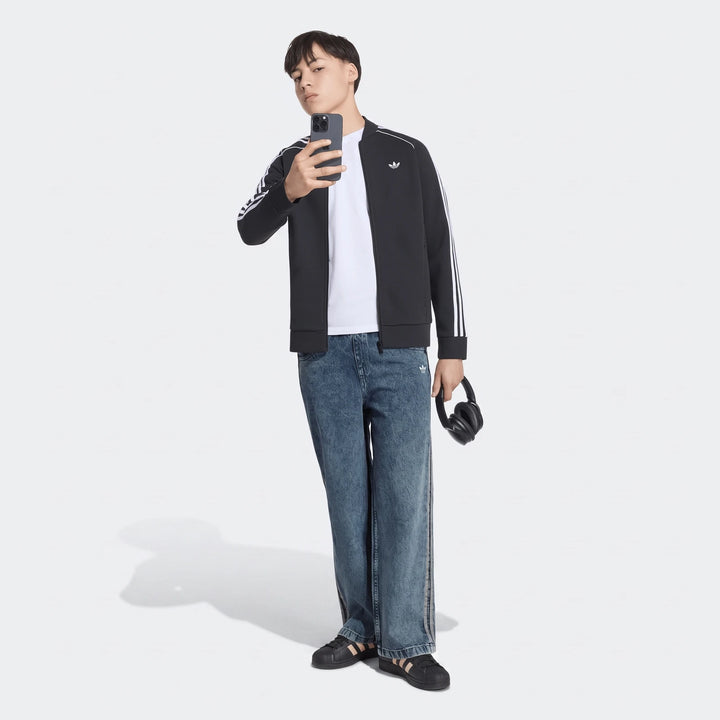 Young man wearing Adidas black track jacket and blue Firebird loose denim track pants, holding a phone and headphones,