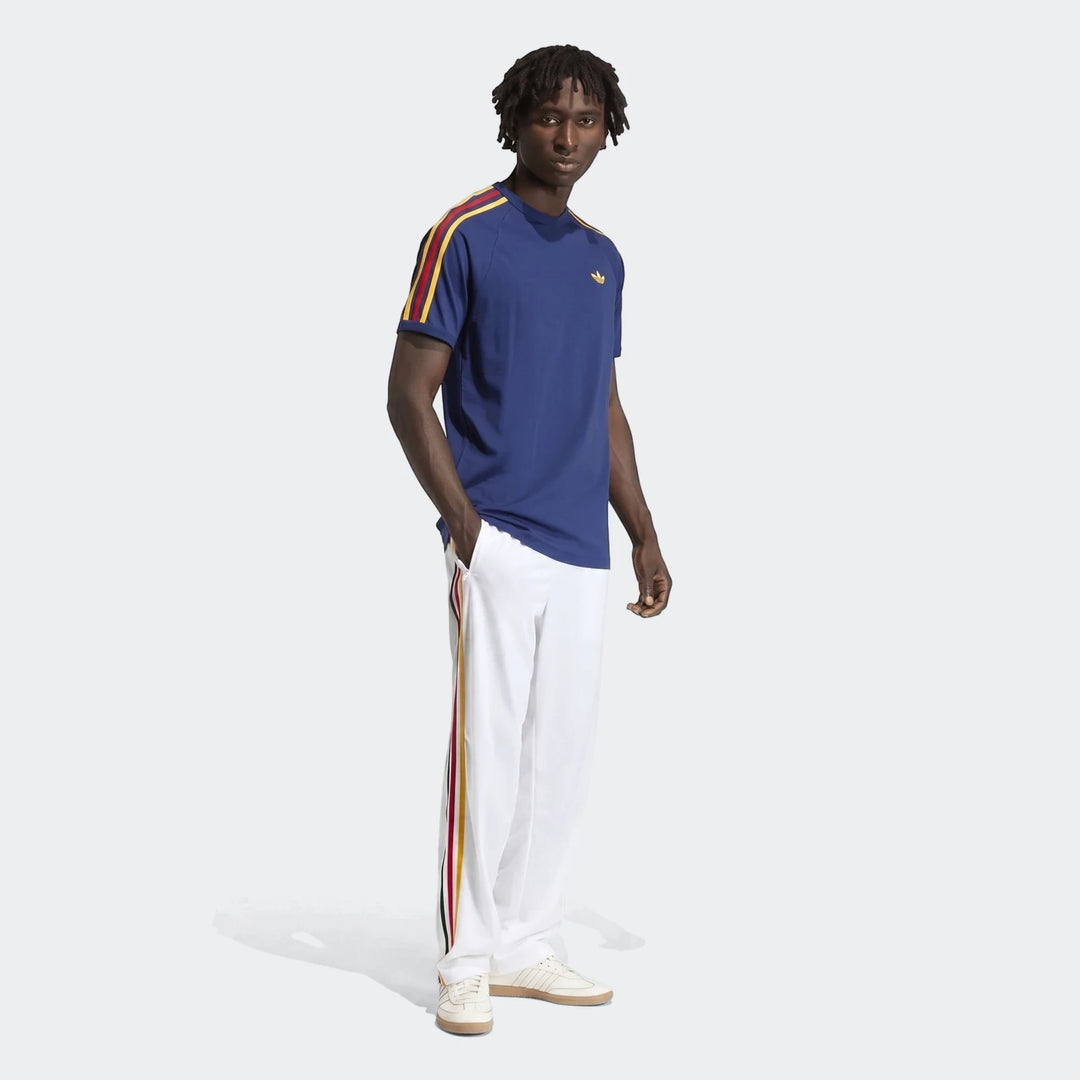 Male model wearing white Adidas Firebird Track Pants with red, yellow, and navy stripes, a navy blue t-shirt, and white
