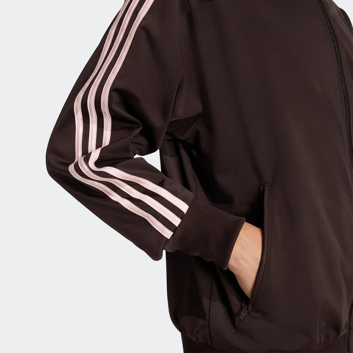 Close-up of a dark brown Adidas Firebird Track Top with light pink stripes on the sleeve, ribbed cuff, and a hand in the