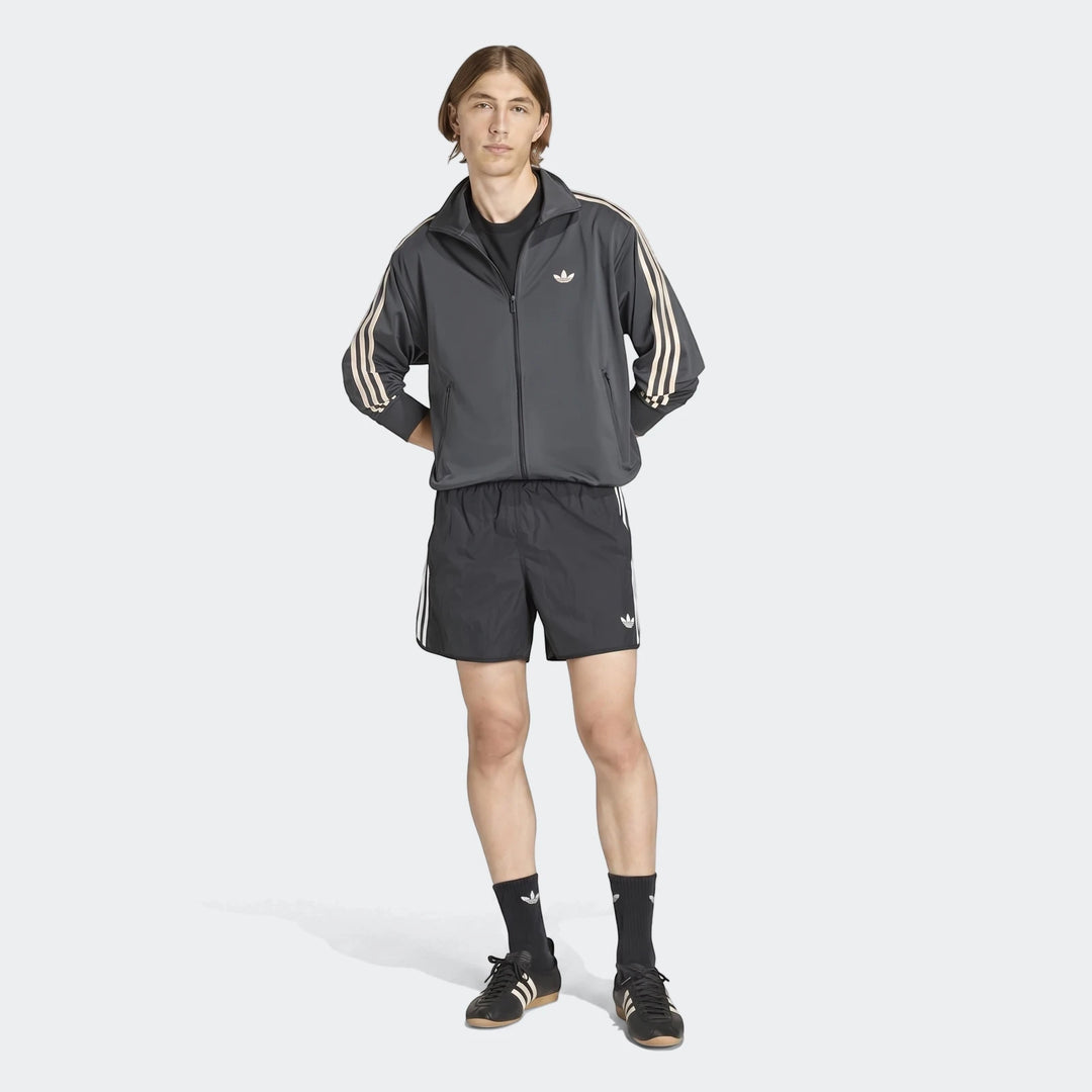 Male model wearing a dark gray Adidas Firebird Track Top with off-white stripes, black shorts, and sneakers, full body shot.