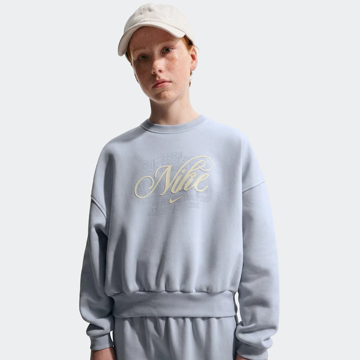 Female model wearing a light blue Nike G Nsw Club Flc Bxy Crew Gfx sweatshirt with a unique script logo.