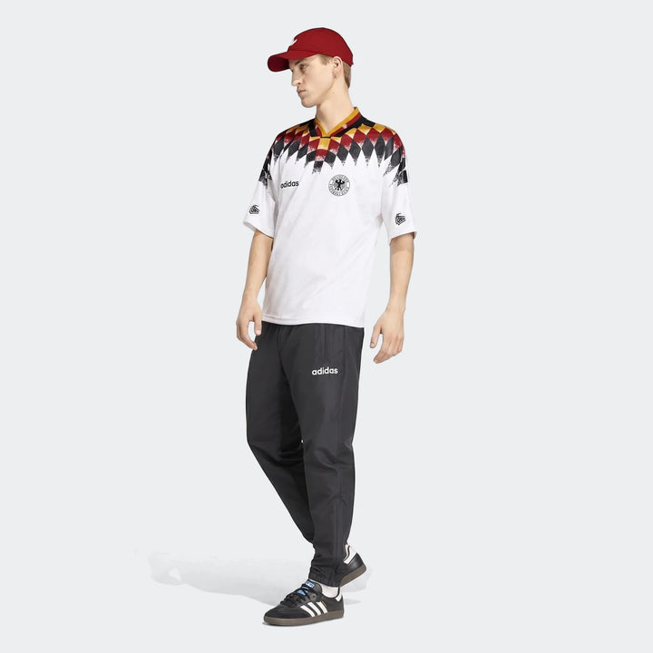Male model wearing a white Adidas Germany 1994 Retro Home Jersey, black track pants, and a red cap, full body shot.