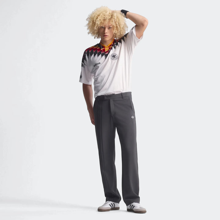 Male model wearing a Germany 1994 Retro Home Jersey, white with a geometric pattern, dark grey trousers, and sneakers.