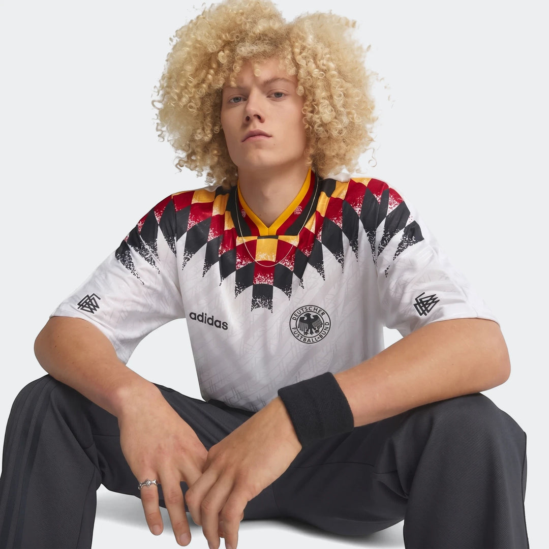A male model wears the white Germany 1994 Retro Home Jersey, featuring black, red, and yellow geometric patterns on the