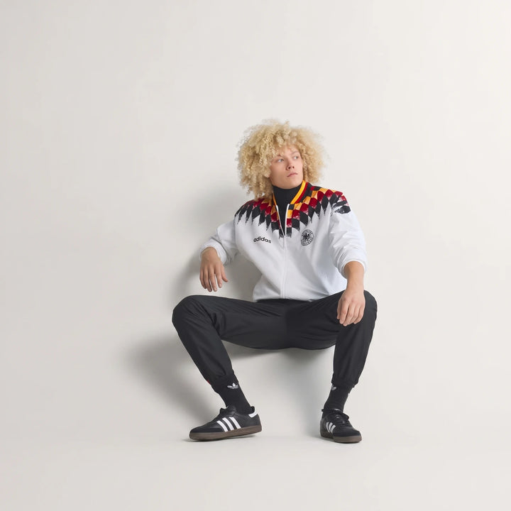 Male model wearing a white, red, yellow, and black Germany 1994 Retro Track Top, paired with black track pants and sneakers,