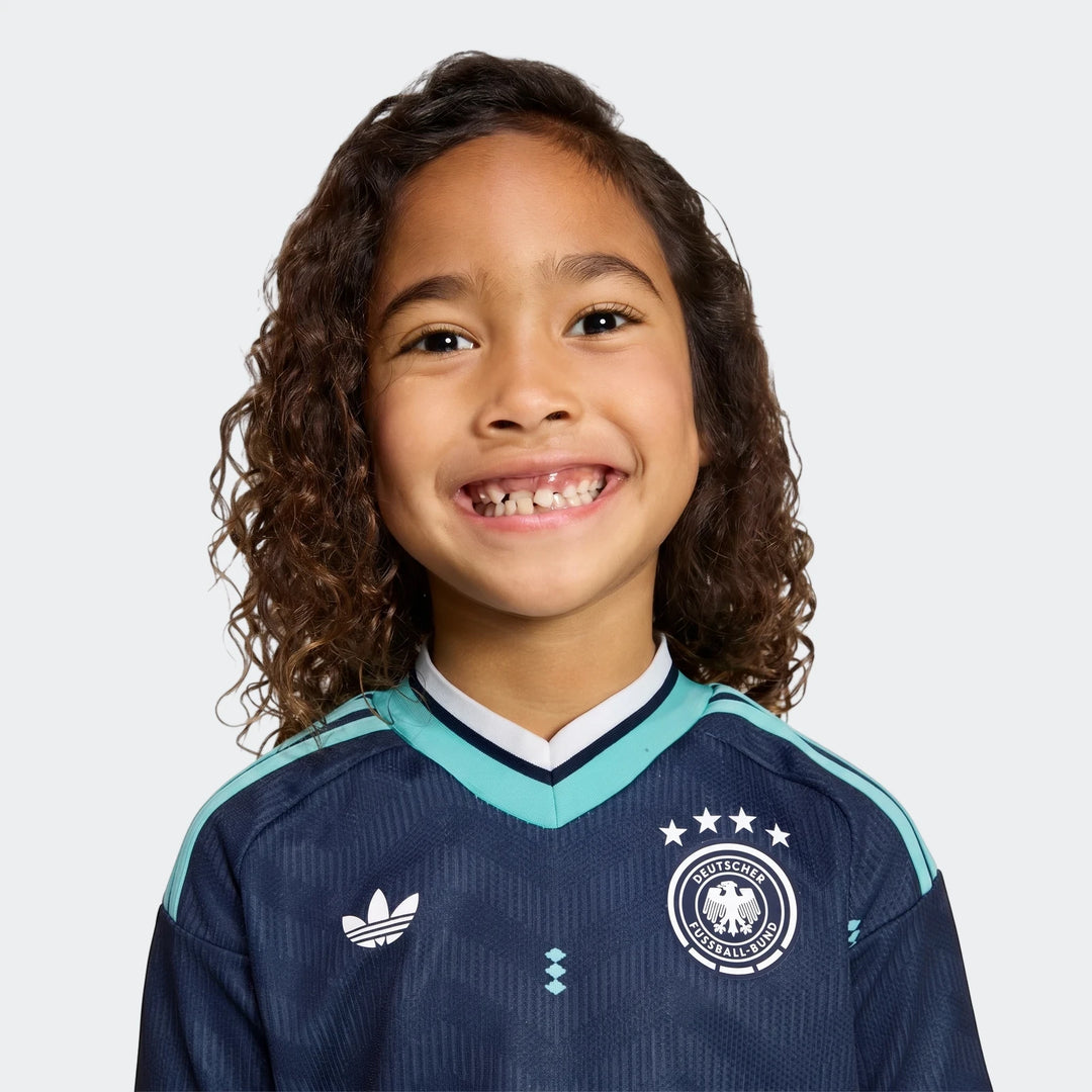Young child smiling in the Germany 25/26 Away Mini Kit, dark blue and teal soccer jersey with team logos.