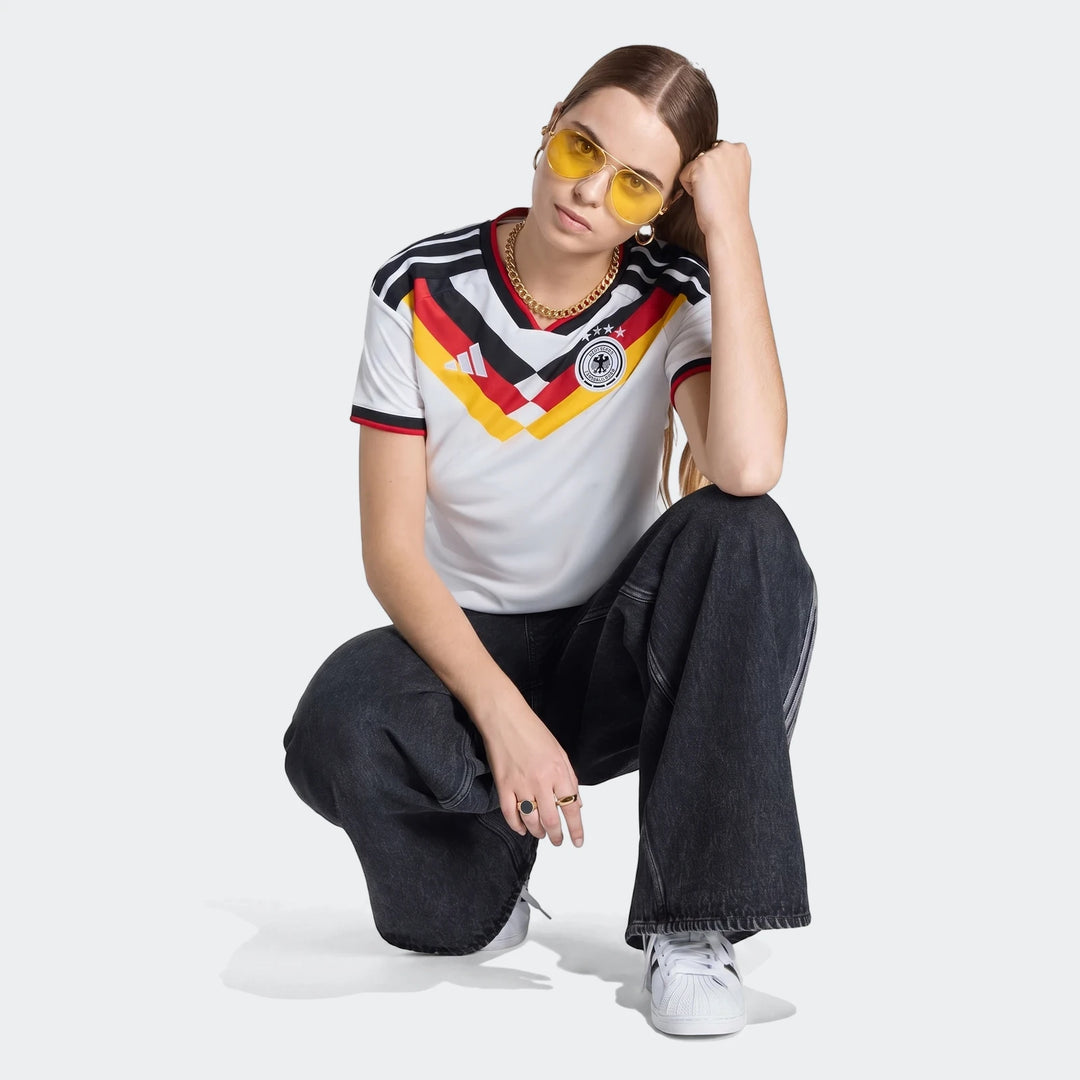 A young woman wearing a white Adidas Germany 25/26 Home Jersey with black, red, and yellow stripes, dark jeans, and white