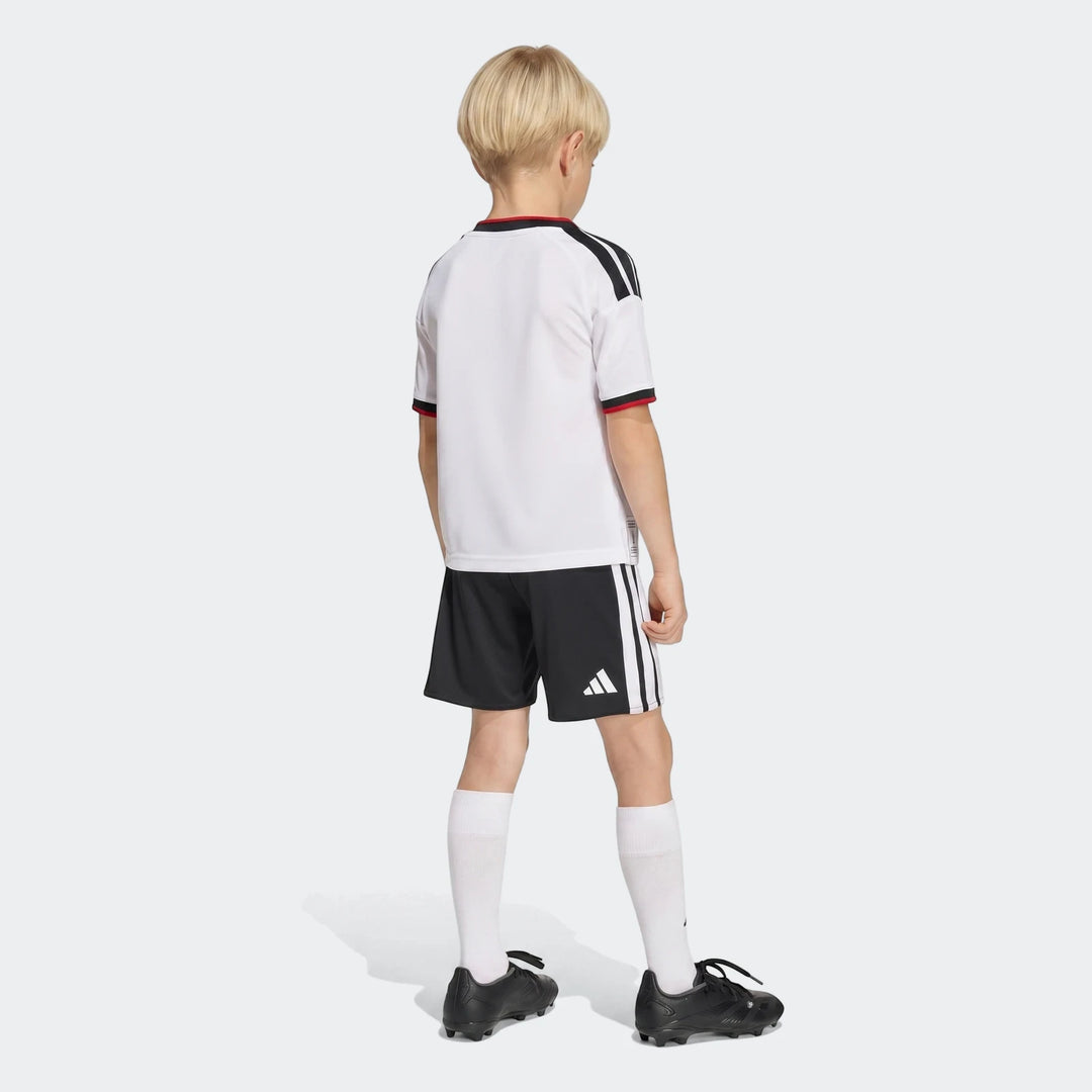 Young boy in white Germany 25/26 Home Mini Kit, black shorts, and cleats, viewed from the back.