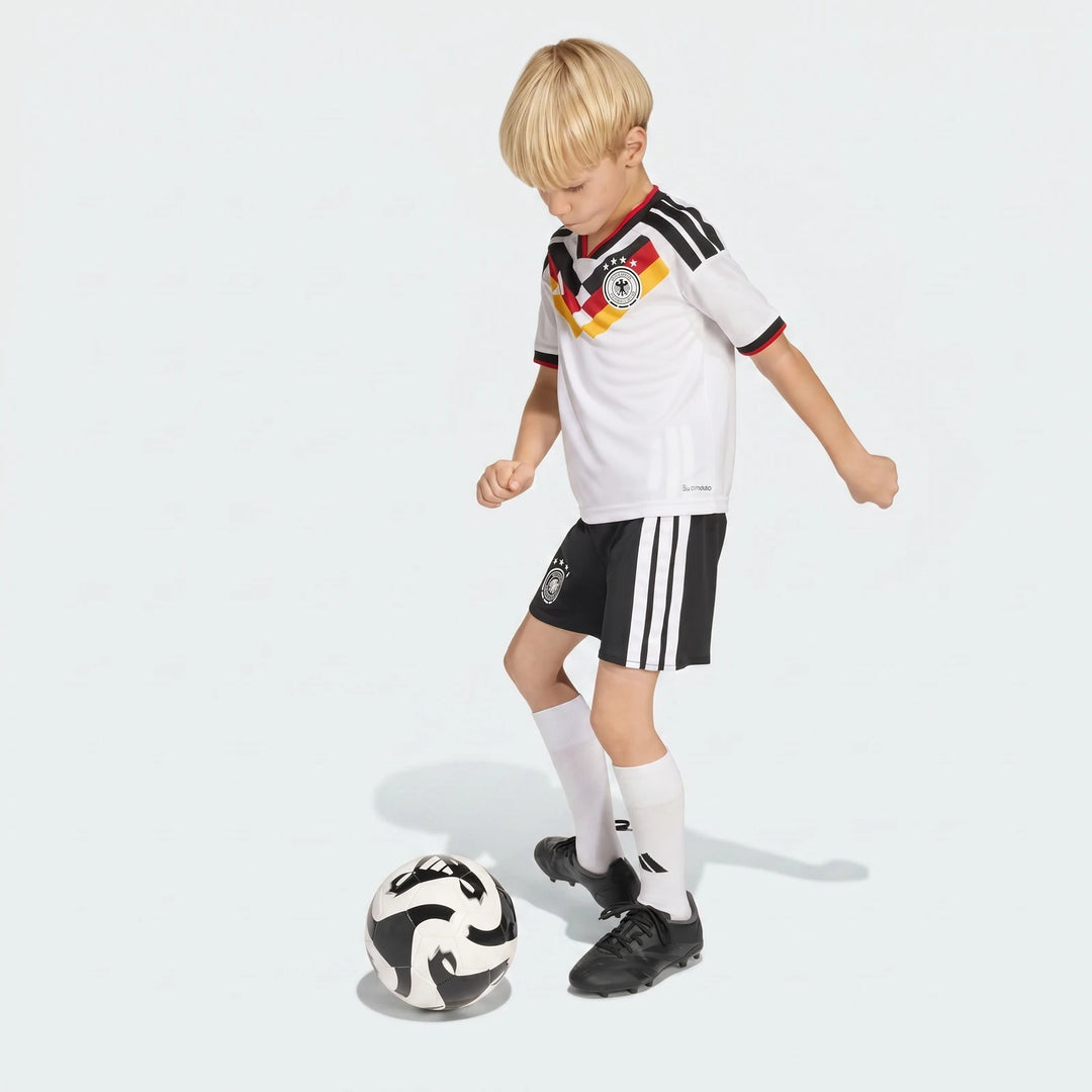 Young boy wearing a Germany 25/26 Home Mini Kit, featuring a white jersey and black shorts, playing with a soccer ball.