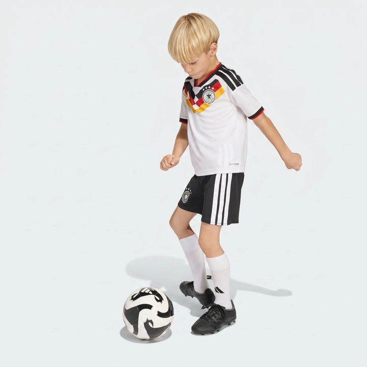 Young boy wearing a Germany 25/26 Home Mini Kit, featuring a white jersey and black shorts, playing with a soccer ball.