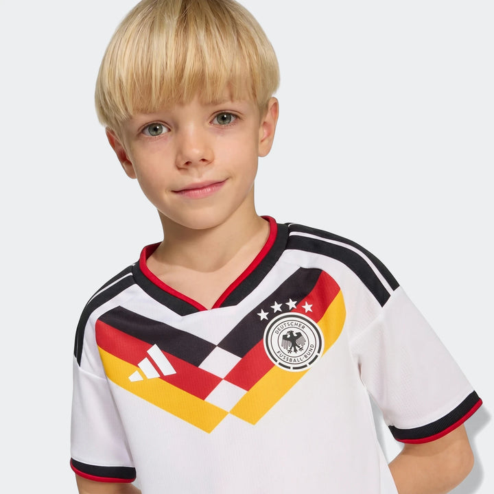 Young boy wearing a white Germany 25/26 Home Mini Kit football jersey with black, red, and yellow design.