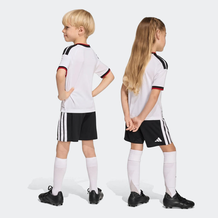 Two children modeling the Germany 25/26 Home Mini Kit, featuring white short-sleeve jerseys and black shorts, shown from the