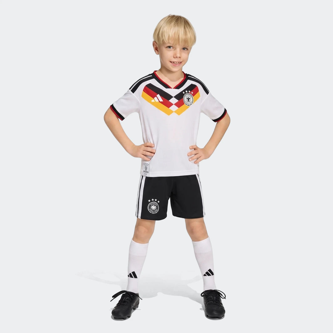 Young boy wearing a white Germany 25/26 Home Mini Kit with black shorts, white socks, and black cleats.