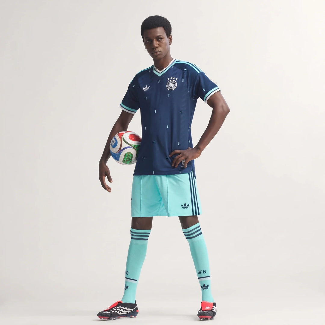 Male model wearing dark blue Adidas Germany 26 Away Authentic Jersey, light blue shorts, and holding a soccer ball.