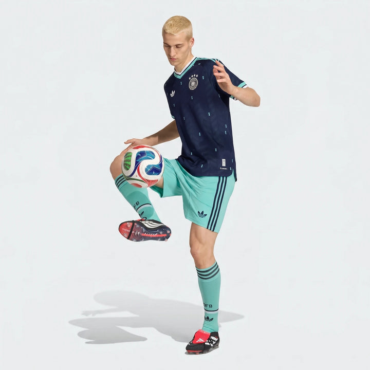 Male model wearing the Germany 26 Away Authentic Jersey, dark blue with light blue details, balancing a soccer ball.