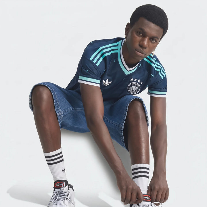 A man wearing the dark blue Adidas Germany 26 Away Authentic Jersey, denim shorts, and white socks, sitting and tying his