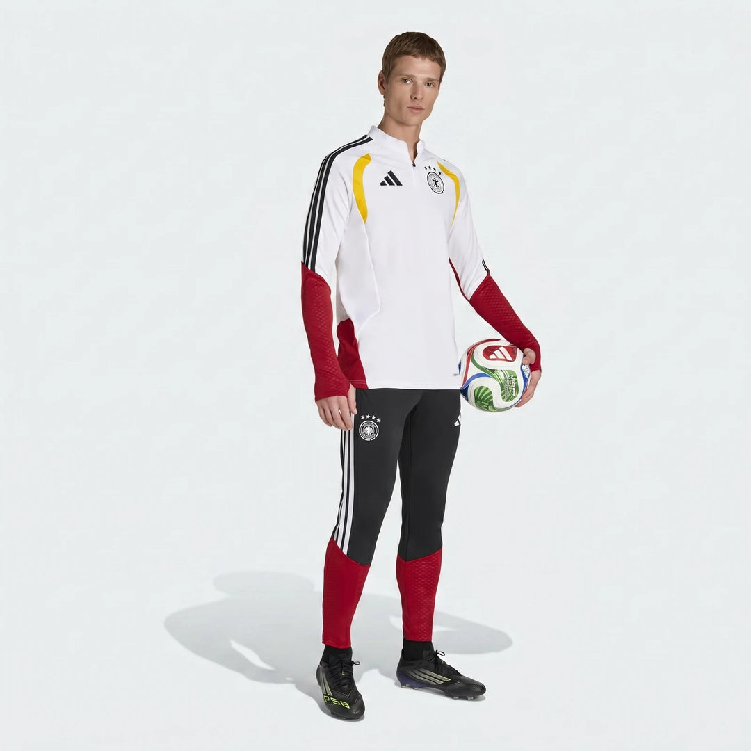A male model wears a white, red, and black Adidas Germany 26 Tiro Training Top and black track pants, holding a soccer ball.