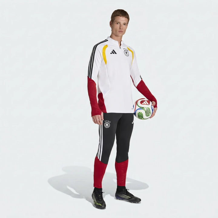 A male model wears a white, red, and black Adidas Germany 26 Tiro Training Top and black track pants, holding a soccer ball.