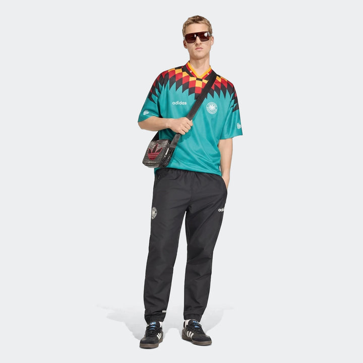 Model wearing a teal and patterned Adidas Germany Away Jersey 1994, black track pants, and black sneakers, with sunglasses