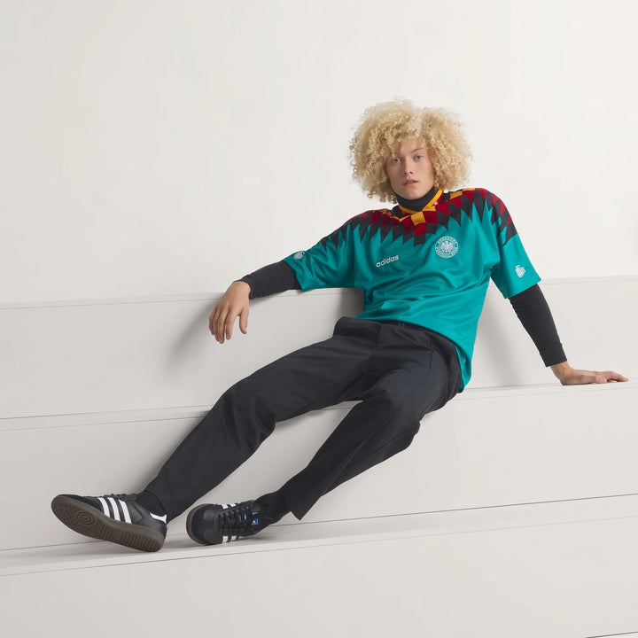Male model wearing a teal Germany Away Jersey 1994 with black, red, and yellow geometric patterns, black pants, and sneakers.
